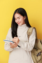 happy asian girl student with backpack writing in a notepad while standing with books isolated over yellow background