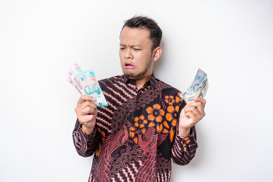 A confused young Asian man is wearing batik shirt and holding cash money in Indonesian rupiah isolated by white background
