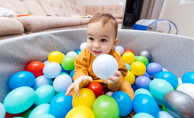 Fototapeta na wymiar Happy child playing at colorful plastic balls. Cute baby boy having fun in ball pit. Activity toys for little kid.