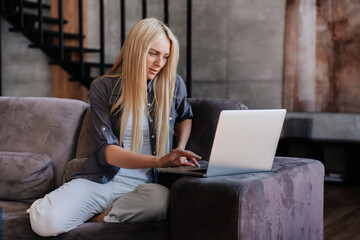 Exhausted blonde caucasian woman in grey shirt and white pants sitting on sofa with laptop remote working at home,, overloaded businesswoman typing on computer. Fatigue concept. Tired Swedish female.