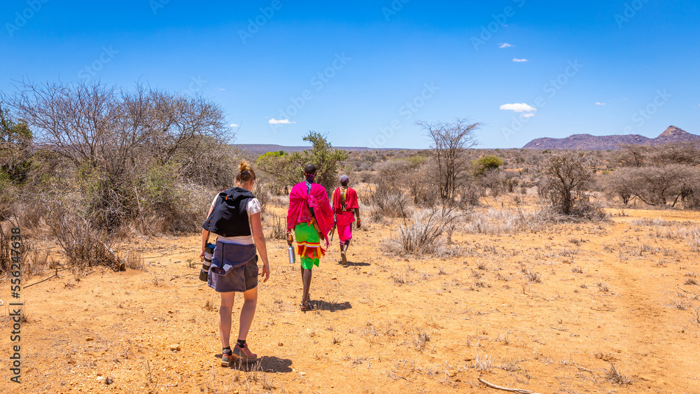 Wall mural maasai tribe men wearing traditional clothes and a tourist walking away in an open kenya africa fiel