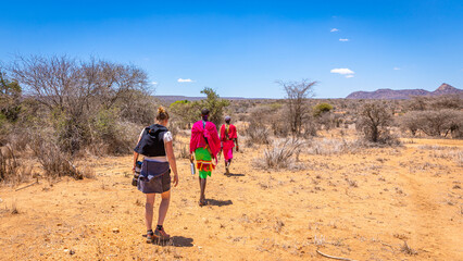 Maasai tribe men wearing traditional clothes and a tourist walking away in an open Kenya Africa field with open blue sky, Laikipia, Kenya.