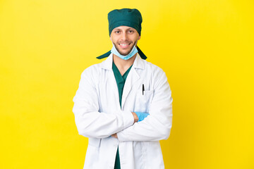 Surgeon blonde man in green uniform isolated on yellow background keeping the arms crossed in...