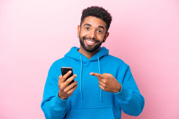 Young Brazilian man isolated on pink background using mobile phone and pointing it