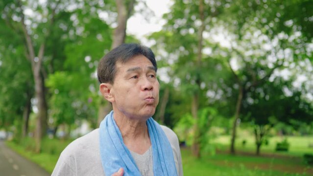 Athlete pensioner drinking water, wiping sweat after outside training, active fit asian mid age man cool down, holding plastic water bottle. Walking under the trees at park garden. Body Hydration