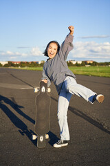 Vertical shot of asian girl feeling excited, skating on longboard, jumping and posing with...