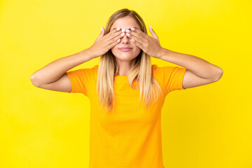 Blonde Uruguayan girl isolated on yellow background covering eyes by hands and smiling
