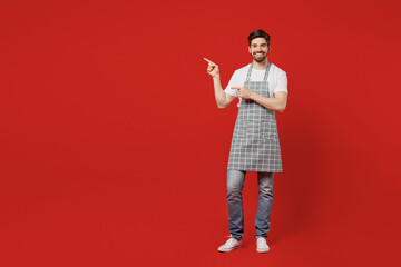 Full body smiling young male housewife housekeeper chef cook baker man wear grey apron point index finger aside on workspace area mock up isolated on plain red background studio. Cooking food concept.