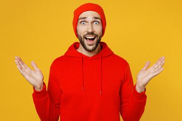 Young excited surprised stunned shocked astonished caucasian man wear red hoody hat look camera spread hands say wow isolated on plain yellow color background studio portrait People lifestyle concept