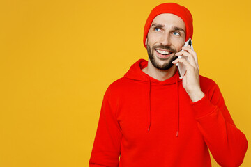 Young smiling fun caucasian man wear red hoody hat talk speak on mobile cell phone conducting pleasant conversation isolated on plain yellow color background studio portrait. People lifestyle concept.