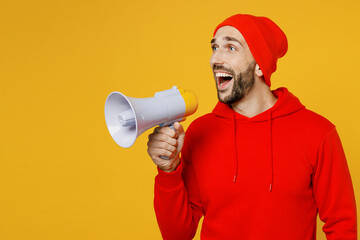 Surprised young shocked excited man wearing red hoody hat hold megaphone scream announces discounts sale Hurry up isolated on plain yellow color background studio portrait. People lifestyle concept
