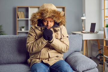 Young man freezing indoors without central heating. African American man wearing warm winter clothes sitting on sofa in very cold room at home looking at camera with angry disappointed face expression