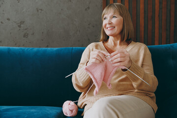 Elderly happy minded woman 50s years old wear casual clothes sits on blue sofa knitting handmade...