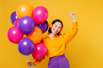 Happy fun young cheerful woman wears casual clothes headphones listen to music raise up hand dance celebrating hold balloons isolated on plain yellow background. Birthday 8 14 holiday party concept.