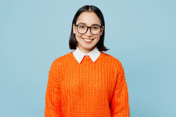 Young satisfied smiling happy fun optimistic smart woman of Asian ethnicity wear orange sweater glasses look camera isolated on plain pastel light blue cyan background studio People lifestyle concept