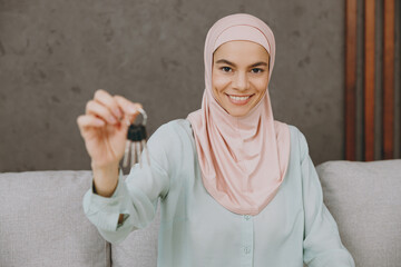 Young calm muslim woman wear hijab casual clothes hold bunch of kes sits on sofa couch stay at home flat rest relax spend free spare time in living room indoor People middle eastern uae islam concept - obrazy, fototapety, plakaty