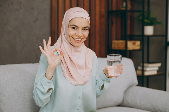 Young muslim woman wears hijab casual clothes drink water show ok sits on sofa couch stay at home flat rest relax spend free spare time in living room indoor. People middle eastern uae islam concept.