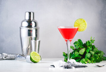 Cosmopolitan classic alcoholic cocktail with vodka, liqueur, cranberry juice, lime, ice. Gray table background, bar tools, copy space