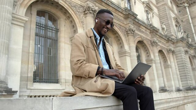 A handsome young African American works in the city on a laptop and communicates with headphones
