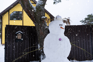 Funny snowman in a hat and scarf on the background of a yellow house in the yard. Winter, winter...