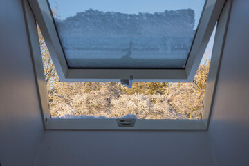 View from snow covered dormer window on roof of house to forest trees covered with snow on frosty sunny winter day. Sweden.