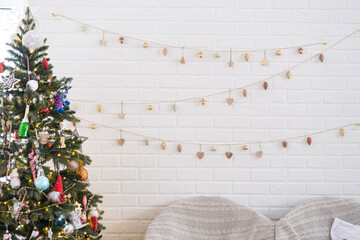 Christmas tree in white interior of a house with loft-style brick walls with garlands of glass toys on a rope. Glowing fairy lights decoration of the studio room. Cozy home in christmas and new year