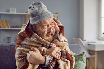 Senior man freezing at home in winter. Elderly man wearing hat and wrapped in blanket sitting on...