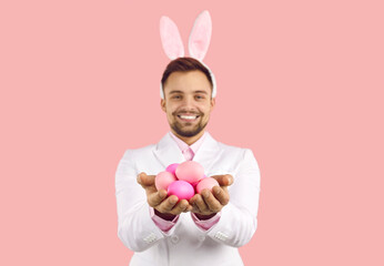 Smiling man wearing bunny ears holding Easter eggs isolated on pastel pink background. Close up of Easter eggs in hands of young Caucasian man wearing white suit and fluffy rabbit ears on his head.