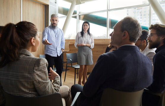 Colleagues Old Man And Young Woman Making Presentation In Office For Group Of People. They Discuss Effective Methods Of Work And Cooperation, Interaction In A Team In Company. Office Meeting Concept.