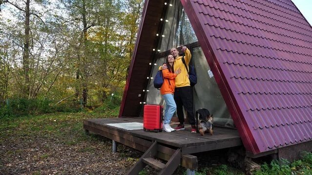 A man and a woman with suitcases are hugging on the porch of a triangular wooden house in the woods. The red tiled roof descends to the ground. Concept of glamping and renting a chalet for the weekend