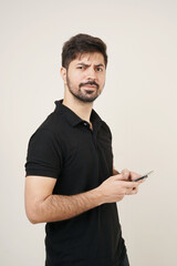 young Asian man holding mobile phone and giving sceptical expression. man in black looking in the camera with sceptical expression holding a smartphone with plain beige background. 