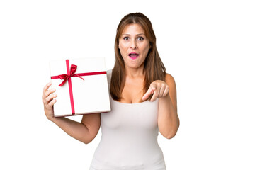Middle age caucasian woman holding a gift over isolated background surprised and pointing front