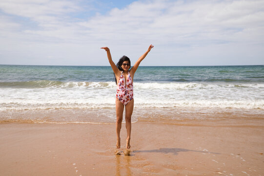 Beautiful and young latin woman is walking along the shore of the beach and posing for photos, while raising her arms and jumping as a symbol of freedom and well-being. Holiday and travel concept.