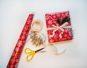 Christmas wrapped rectangle gift and decorative elements on white table; red paper, yellow scissors, gold stars and white christmas ribbon on white background