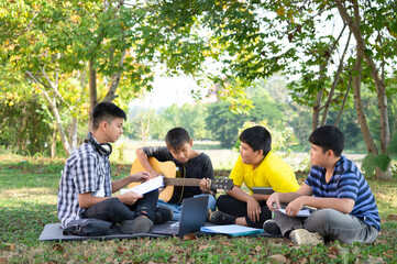 asian teenagers boys are sitting, practicing, playing guitar together on weekend, four of friends having fun in the garden