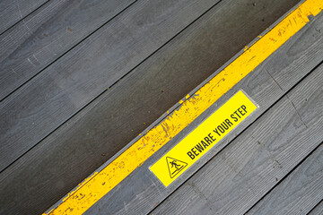 "Beware you step" caution icon symbol with yellow line at the edge of difference level on wooden panel floor. Safety sign in transportation mode object photo, close-up/