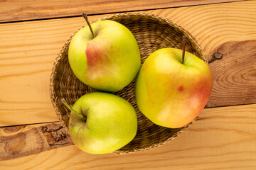Three sweet green apples in a straw plate on a wooden table, macro, top view.