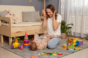 Portrait of smiling happy attractive dark haired woman playing with her infant baby, posing on carpet, having fun with toddler daughter, being photographed among colorful toys.