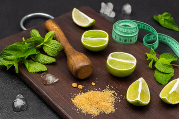 Lime halves, mint sprigs, wooden pestle and measuring tape on a board. Pieces of ice on the table.
