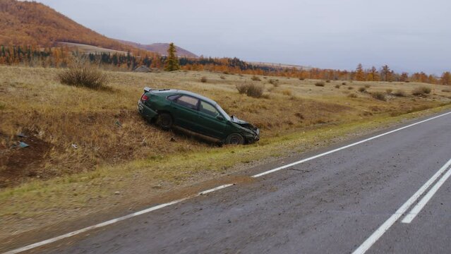 Broken emerald-colored car stands on side of road in valley with ridges. Automobile after traffic accident, unsuitable for further use, is located near mountain roadway. Highway crash