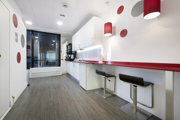 Kitchen of a coworking office with a red countertop and white cabinets, stainless steel stools with...