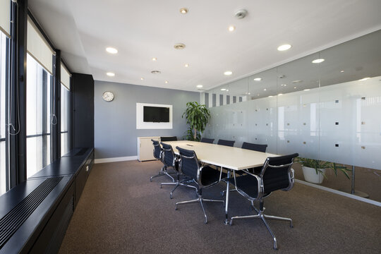Boardroom with long wooden table with black swivel chairs and wall of windows, a large plant, glass partition, carpeted floors and flat tv on the wall