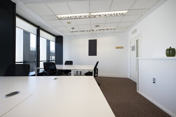 Empty white shared office desks in an office with carpeted floors, large windows with roller blinds and technical ceilings