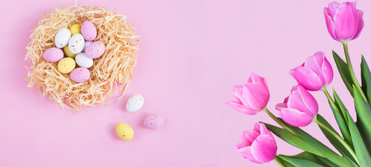 Holiday background. Colored easter eggs in a nest of straw and tulips on the pink background. Copy space.