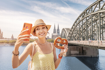 Happy tourist girl taking selfie photo with a pretzel and Cologne cathedral building and bridge...