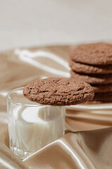 Brown american cookie closeup with a glass of milk on beige silk fabric background. Minimal style. Bakery and pastry concept. Idea for breakfast.