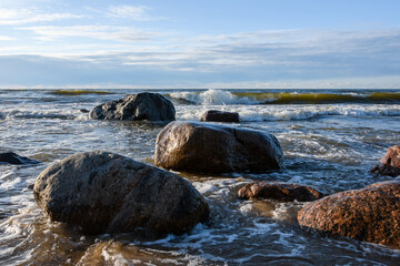 Sea waves roll and break on the coastal granite rocks. Sunny spring evening, Narva Bay, Baltic Sea