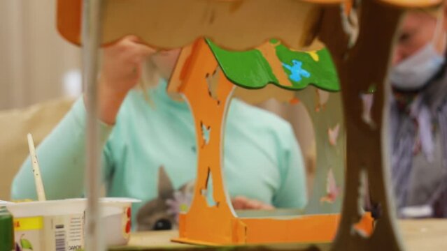 The caucasian child in a turquoise sweater paints a homemade bird house with a brush against the background of a colorful painted plywood craft, standing at the table