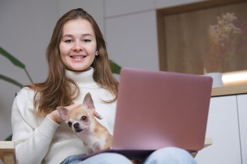 Portrait of young beautiful woman freelancer, teenager girl hug with Chihuahua dog, holding on hands little puppy at home sitting in armchair, smile using laptop computer