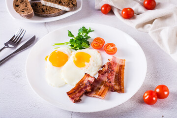 Homemade breakfast fried bacon and eggs, tomatoes and greens on a plate on the table.